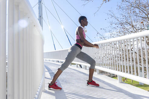 Junge Sportlerin, die sich die Beine vertritt, während sie auf einer Brücke an einem sonnigen Tag übt - RFTF00001