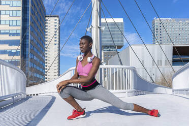 Young woman wearing headphones stretching legs on bridge looking away - RFTF00004