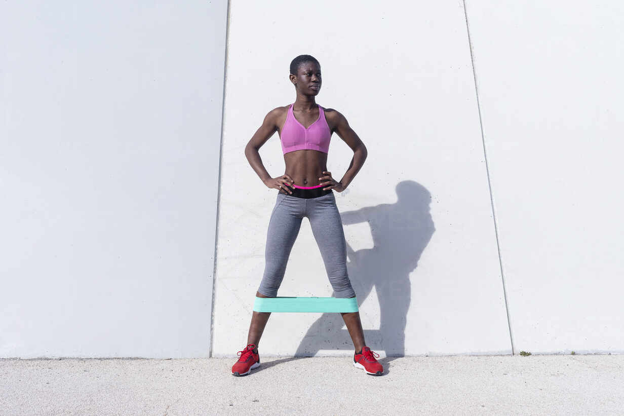 Fit young woman with rubber band doing leg exercise while standing against white wall stock photo