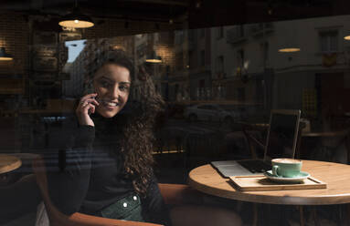 Junge Freiberuflerin, die durch ein Glasfenster mit ihrem Handy telefoniert - DAMF00744