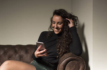 Lächelnde Freiberuflerin, die auf dem Sofa in einem Café sitzt und über ihr Telefon Textnachrichten verschickt - DAMF00748