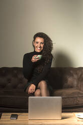 Smiling female entrepreneur holding coffee cup while sitting on sofa in cafe - DAMF00750