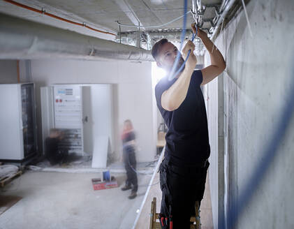 Männlicher Elektriker bei der Installation von Kabeln an der Decke in einer Werkstatt - CVF01669