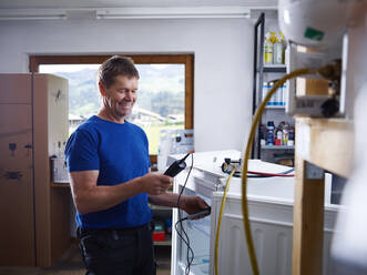 Lächelnder männlicher Elektriker, der ein Gerät hält, während er die Temperatur in einer Werkstatt überprüft - CVF01676