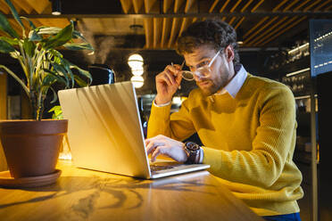 Männlicher Besitzer hält eine Brille in der Hand, während er in einem Café am Laptop arbeitet - VPIF03655