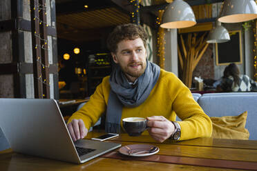 Männlicher Unternehmer schaut weg, während er eine Kaffeetasse am Laptop in einem Café hält - VPIF03674