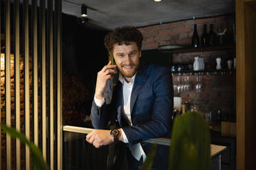 Lächelnder männlicher Unternehmer im Anzug, der am Geländer eines Cafés lehnt und mit seinem Smartphone telefoniert - VPIF03695