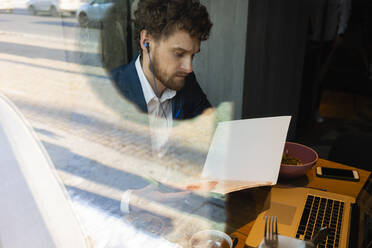 Männlicher Unternehmer, der während eines Videogesprächs auf seinem Laptop im Café sitzend eine Strategie liest - VPIF03699