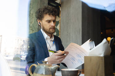 Geschäftsmann mit In-Ear-Kopfhörern, der in einem Café sitzt und seine Strategie überprüft - VPIF03700