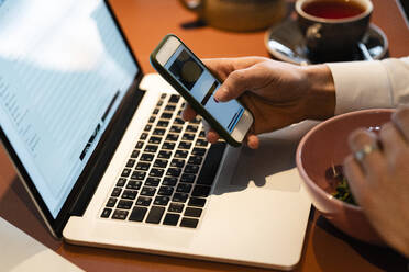 Geschäftsmann, der ein Smartphone über einem Laptop benutzt, während er in einem Café sitzt - VPIF03715