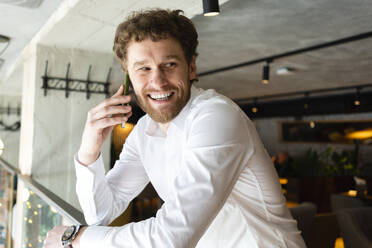 Happy businessman talking on smart phone while leaning on railing at cafe - VPIF03718