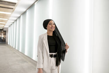 Glückliche arabische Frau, die beim Einkaufen im Einkaufszentrum an der Wand steht - JCCMF01434