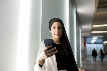 Junge arabische Frau, die ein Smartphone benutzt, während sie in der Nähe einer Wand in einer Einkaufsstraße steht - JCCMF01436
