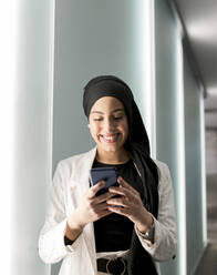 Smiling Arab woman with headscarf using smart phone standing by wall in shopping mall - JCCMF01437