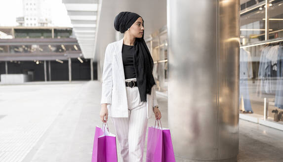 Nachdenkliche arabische Frau mit Kopftuch hält Taschen beim Einkaufen im Einkaufszentrum - JCCMF01442