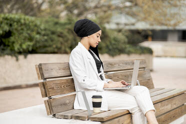 Arabische Geschäftsfrau mit wiederverwendbarem Becher, die auf einer Bank im Freien sitzend am Laptop arbeitet - JCCMF01454