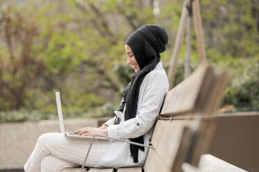Lächelnde Unternehmerin arbeitet an einem Laptop, während sie auf einer Bank im Freien sitzt - JCCMF01458