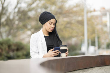 Junge arabische Frau, die ein Smartphone benutzt und einen wiederverwendbaren Becher hält, während sie an einer Stützmauer steht - JCCMF01465