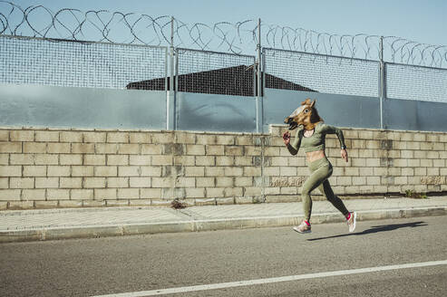 Woman in horse mask running on road during sunny day - JCMF01908