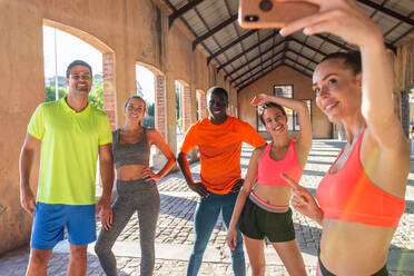 Eine Gruppe verschiedener Läufer in Sportkleidung steht auf der Straße und macht ein Selfie mit dem Smartphone während des Trainings im Sommer - ADSF21635