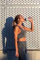 Side view of tired sweaty female runner standing on street on sunny day and enjoying fresh water after active workout - ADSF21643