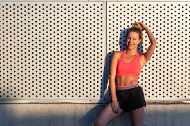 Delighted female athlete in sportswear standing near building in city and relaxing after workout in summer on sunny day - ADSF21644