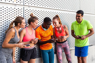 Gruppe gemischtrassiger Läufer in Sportkleidung, die sich nach dem Training in der Stadt an eine Hauswand lehnen und Smartphones benutzen - ADSF21646