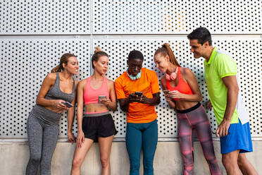 Gruppe gemischtrassiger Läufer in Sportkleidung, die sich nach dem Training in der Stadt an eine Hauswand lehnen und Smartphones benutzen - ADSF21647