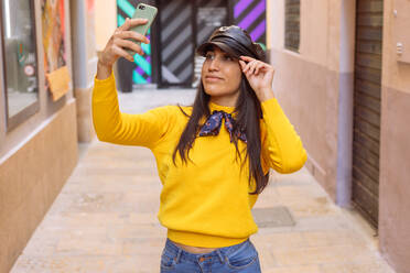 Positive millennial ethnic female in bright yellow sweater and stylish leather cap taking selfie on mobile phone while standing on narrow urban street - ADSF21652