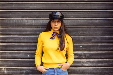 Confident modern young ethnic female in bright yellow sweater and trendy black leather cap looking at camera while standing against gray wall - ADSF21653