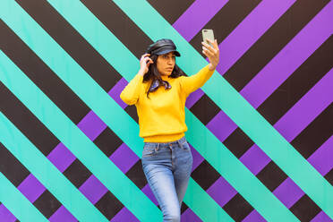 Confident young ethnic female wearing skinny jeans with bright yellow sweater and leather cap taking selfie on mobile phone while standing against colorful striped wall - ADSF21655