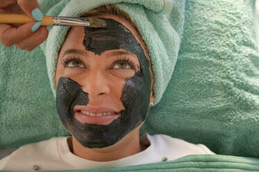 From above of crop anonymous female cosmetologist applying black clay mask on face of happy smiling lady lying on couch with towel on head during skincare procedure - ADSF21665
