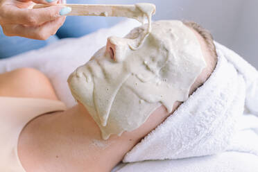 Eine nicht erkennbare Kosmetikerin trägt eine Alginatmaske auf das Gesicht einer anonymen jungen Kundin auf, die mit geschlossenen Augen auf der Couch liegt - ADSF21669