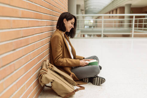Positive Studentin, die auf dem Boden sitzt und ein Buch liest, während sie im Korridor der Universität studiert - ADSF21674
