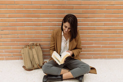 Positive Studentin, die auf dem Boden sitzt und ein Buch liest, während sie im Korridor der Universität studiert - ADSF21675
