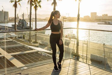 Full body of slim sporty female in activewear looking away while standing on terrace near city waterfront and resting after outdoor workout at sunset time - ADSF21684