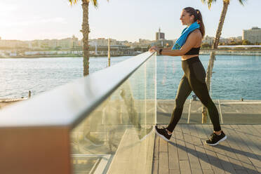 Ganzkörper-Seitenansicht einer fitten Frau in schwarzer Sportkleidung, die ihre Beine beim Fitnesstraining am Ufer der Stadt ausstreckt - ADSF21685