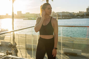 Positiv gestimmte Frau in Sportkleidung, die drahtlose Ohrhörer einstellt, während sie sich auf ein Fitnesstraining im Freien am Stadtkai bei Sonnenuntergang vorbereitet - ADSF21686