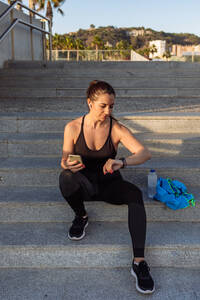 Junge fitte Läuferin in Sportkleidung, die auf einer Treppe sitzt und nach dem Training im Freien ihren Fitness-Tracker und ihr Smartphone überprüft - ADSF21687