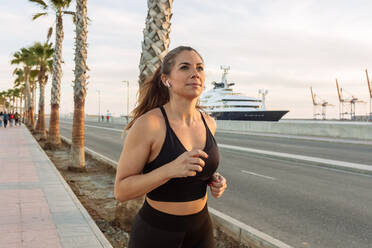 Positiv entschlossene junge sportliche Frau in schwarzer Sportkleidung beim Joggen entlang einer städtischen Böschung während eines Fitness-Trainings bei Sonnenuntergang - ADSF21688