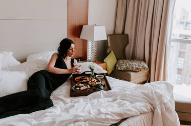 Ruhige Frau im eleganten schwarzen Kleid, die sich auf dem Bett ausruht und verschiedene leckere Speisen auf einem Holztablett in einem Luxushotel genießt - ADSF21724