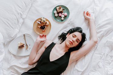 Top view of carefree female lying on bed with closed eyes with assorted tasty desserts and enjoying weekend in luxury hotel - ADSF21725