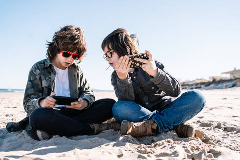 Zwei Kinder spielen mit ihrem Handy am Strand an einem sonnigen Wintertag - ADSF21749