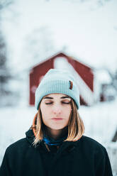Gelassene Frau in warmer Oberbekleidung steht im Winter in einem Dorf und genießt die Natur mit geschlossenen Augen - ADSF21754