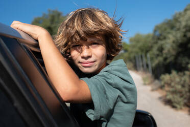 Seitenansicht eines Jungen mit lockigem Haar, der in einem offenen Autofenster sitzt und in die Kamera schaut, während er ein Sommerabenteuer im Gebirge genießt - ADSF21755