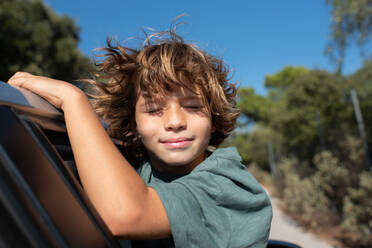 Seitenansicht eines Jungen mit lockigem Haar, der im offenen Fenster eines Autos sitzt und die Augen schließt, während er das Sommerabenteuer in den Bergen genießt - ADSF21756