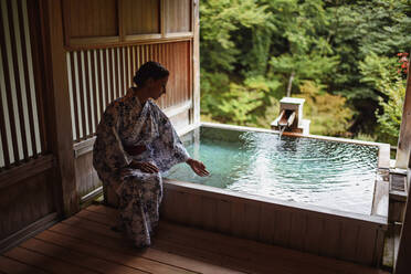 Junge kaukasische Frau in einem Kimono, die das Thermalwasser berührt, bevor sie ein traditionelles Bad in einem japanischen Onsen genießt - ADSF21792