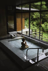 Young caucasian woman kneeling inside a thermal waters bath and meditating with hands in prayer at a traditional Japanese onsen - ADSF21802