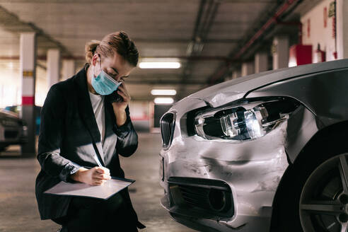 Unerkennbar konzentrierte weibliche Versicherungsangestellte in formellem Anzug und Schutzmaske, die mit einem Mobiltelefon spricht, während sie nach einem Unfall die Schäden am Auto begutachtet und den Antrag ausfüllt - ADSF21812