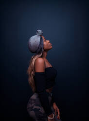 Seitenansicht eines attraktiven jungen afrikanischen weiblichen Modells in stilvollem Outfit mit nackten Schultern und traditioneller Kopfbedeckung, das vor einem dunklen Hintergrund steht und wegschaut - ADSF21826